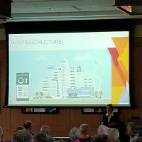 A woman speaks to a crowd in front of a presentation that has the word "Infrastructure" as a title